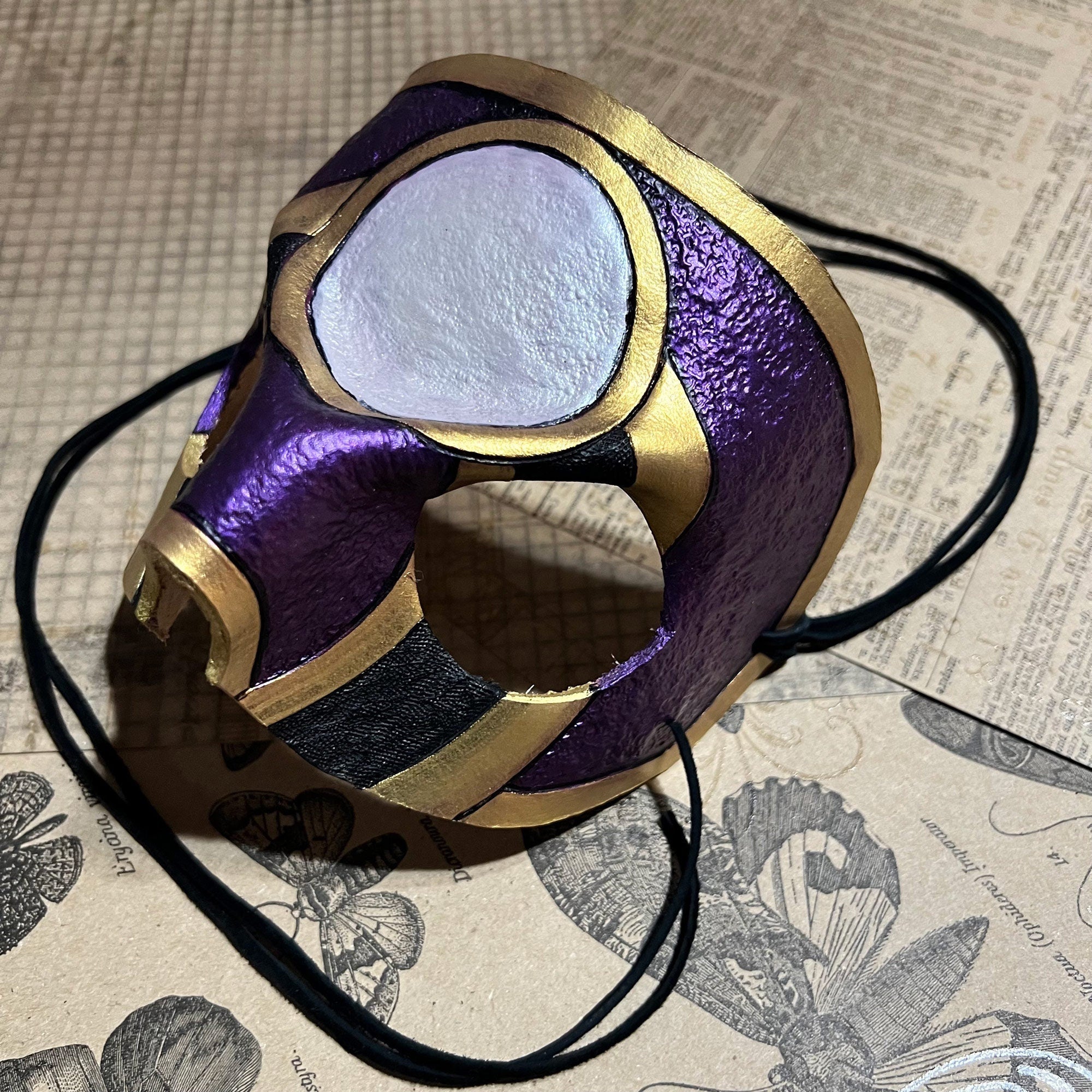 A purple and gold masquerade mask with a black strap, resting on a vintage-style illustrated paper background.
