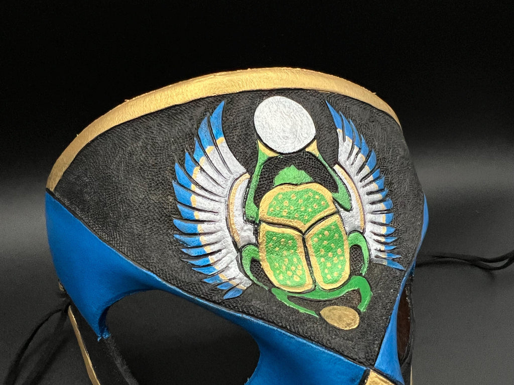 A close-up of a handcrafted mask featuring a green scarab beetle with gold accents, white and blue wings, and a black background, with blue and gold textured panels.