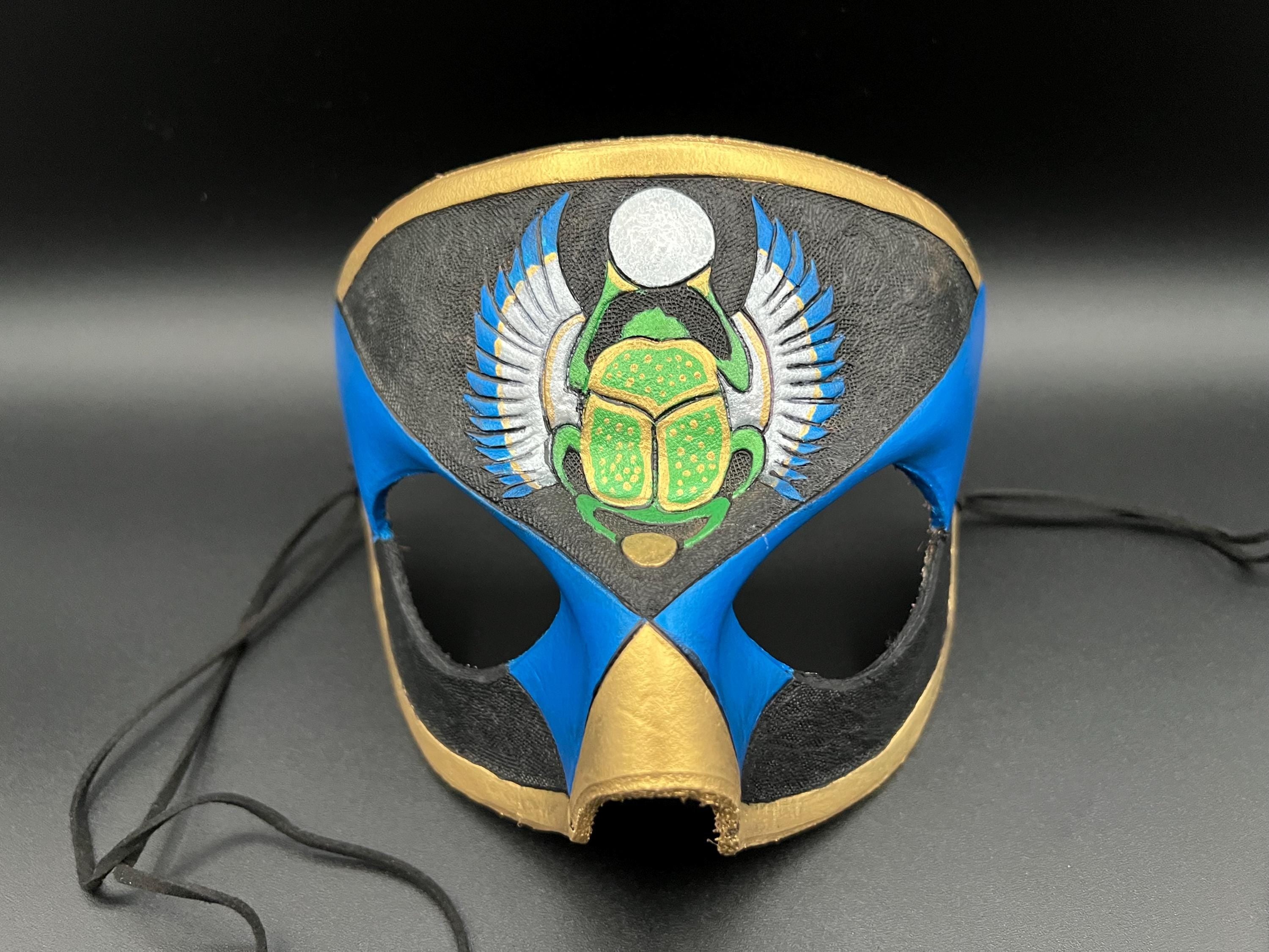 A leather mask featuring a blue and black design with gold trim, a green scarab beetle motif, and silver wings, shown on a dark gray background with black ties