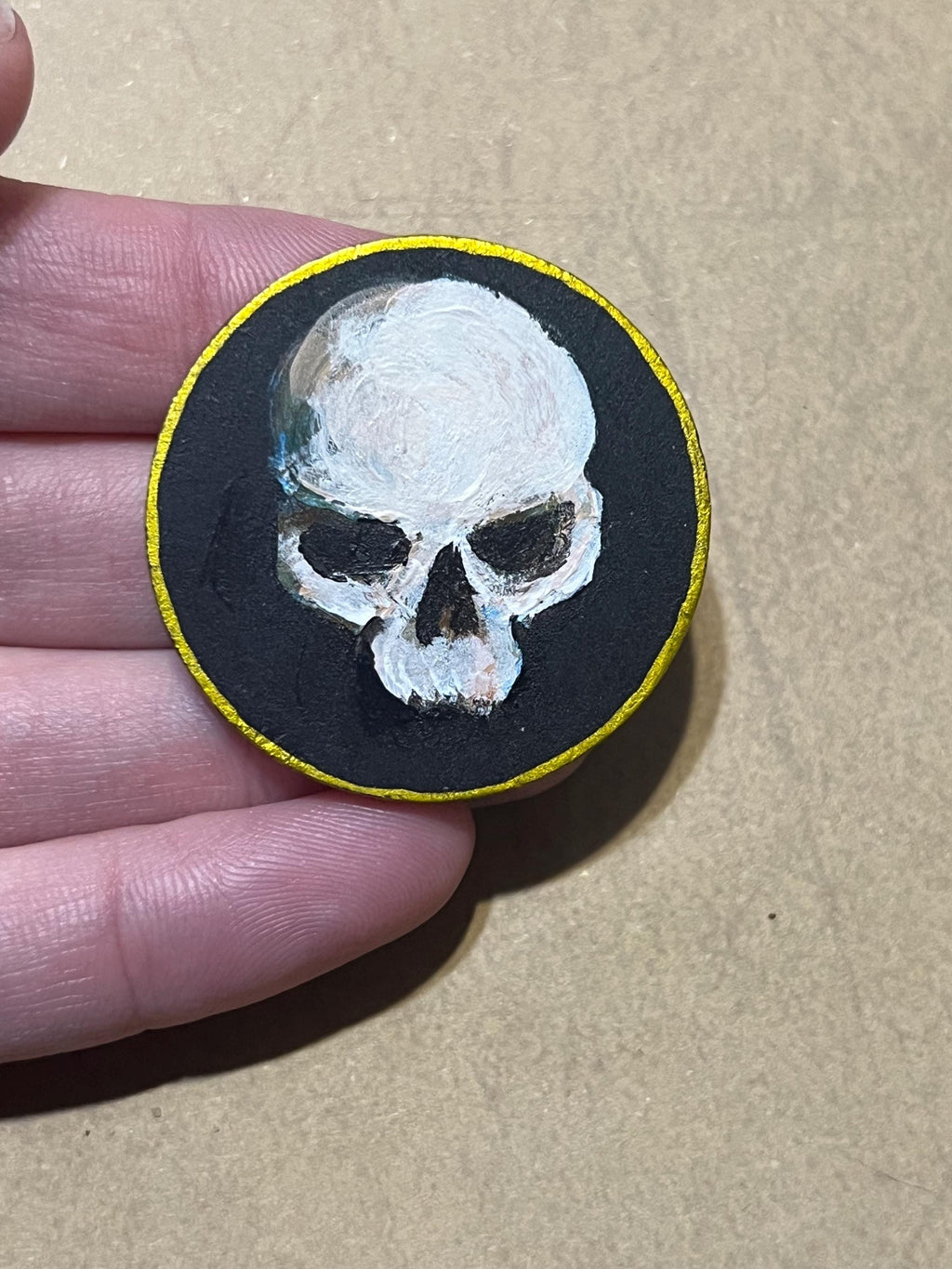 A hand holding a round black patch featuring a white skull painted on a black background with a thin yellow border, against a tan cardboard-like surface.