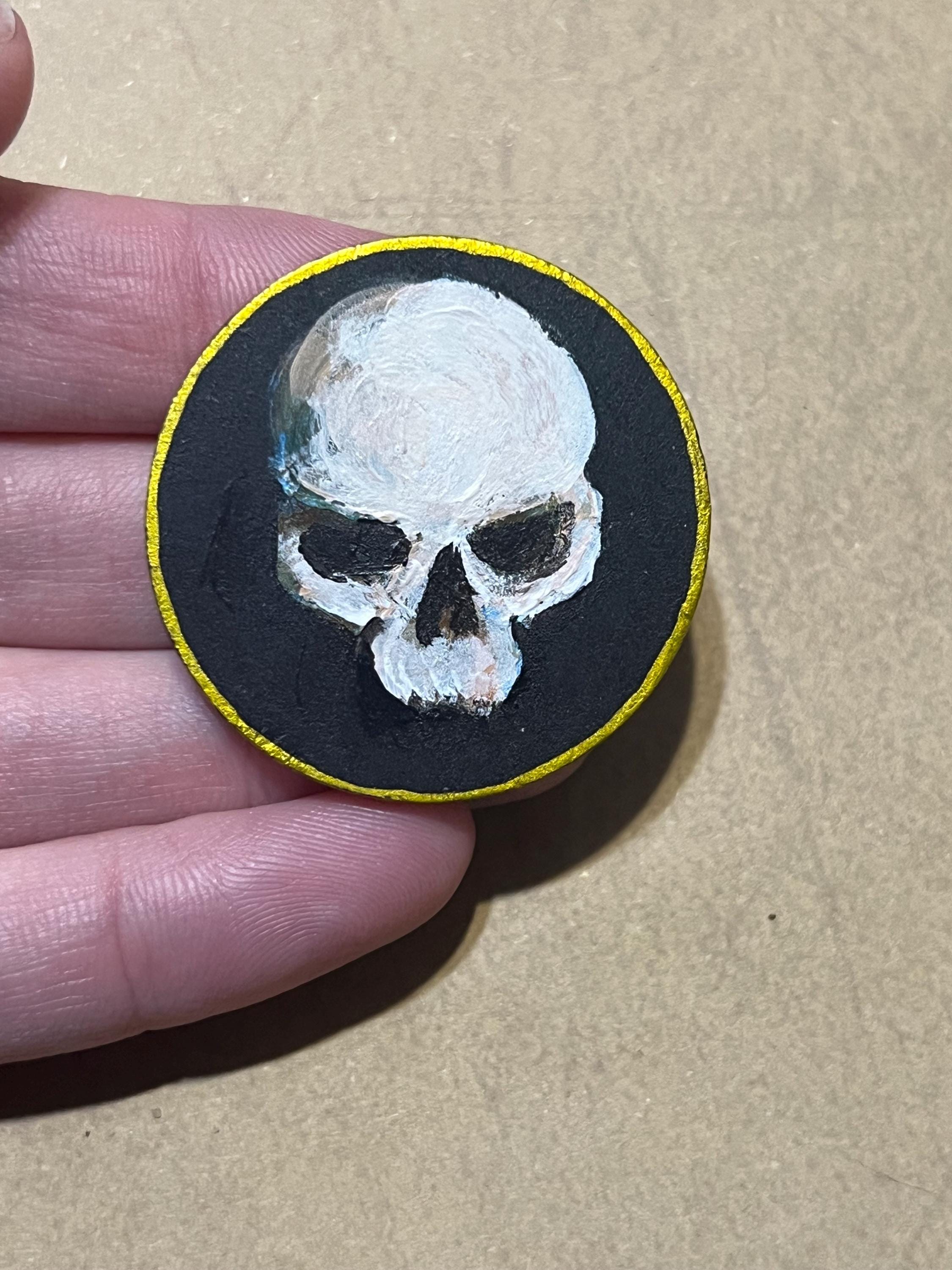 A hand holding a round black patch featuring a white skull painted on a black background with a thin yellow border, against a tan cardboard-like surface.