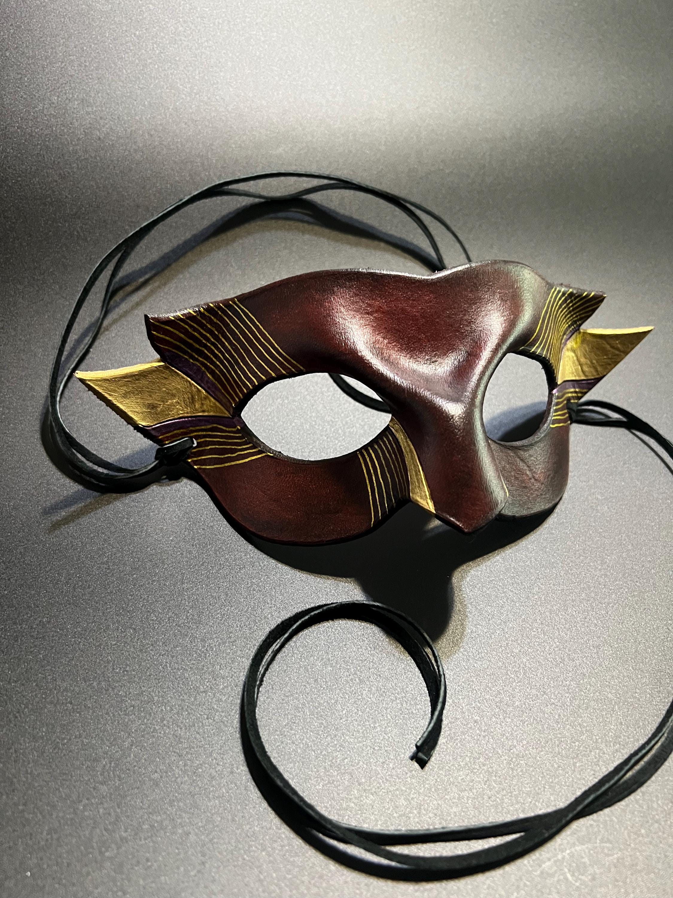 A decorative masquerade mask with a dark reddish-brown center, gold patterned side wings, and black suede ties, photographed on a gray studio background.