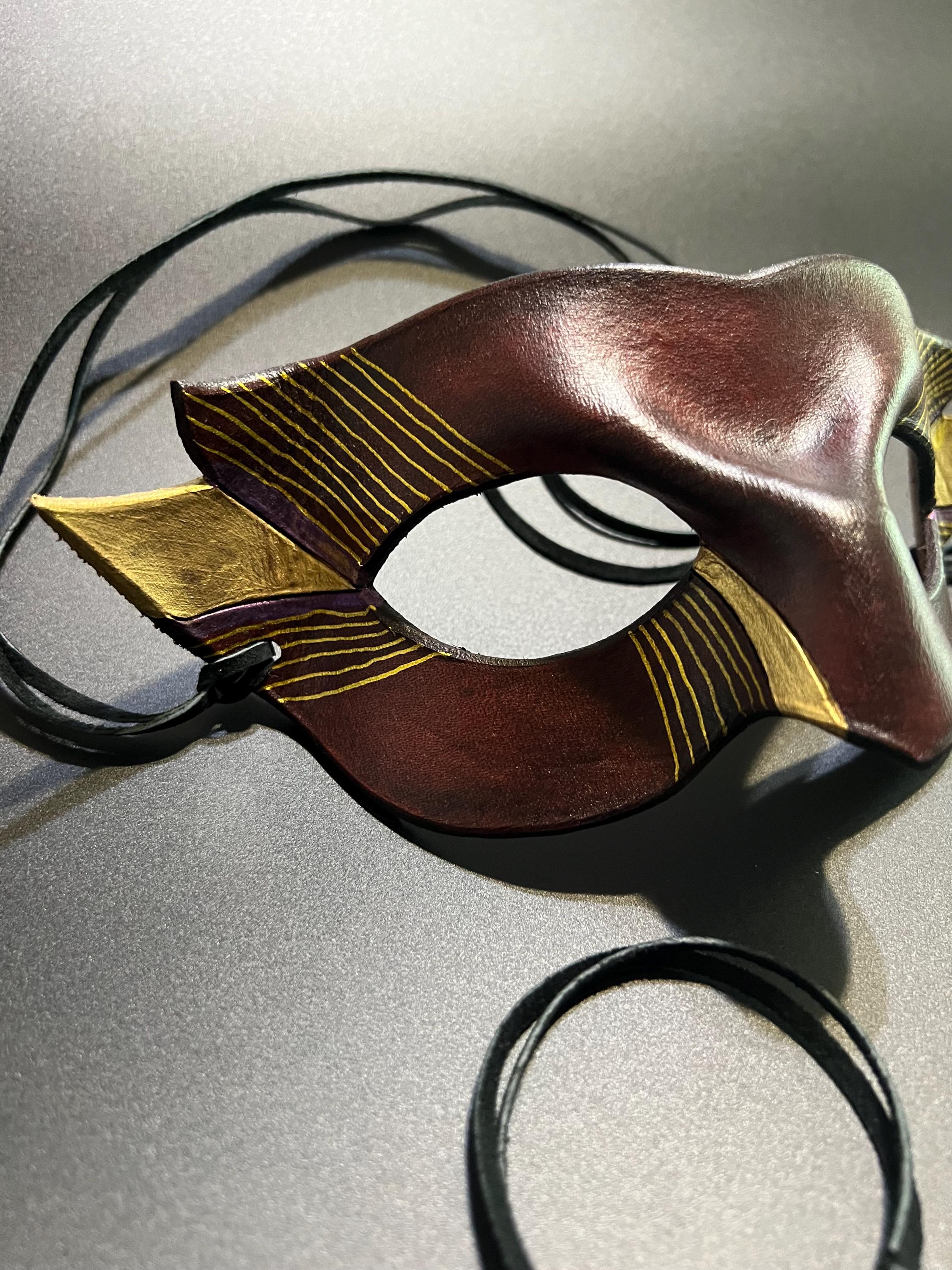 A decorative masquerade mask with a dark reddish-brown center, gold patterned side wings, and black suede ties, photographed on a gray studio background.