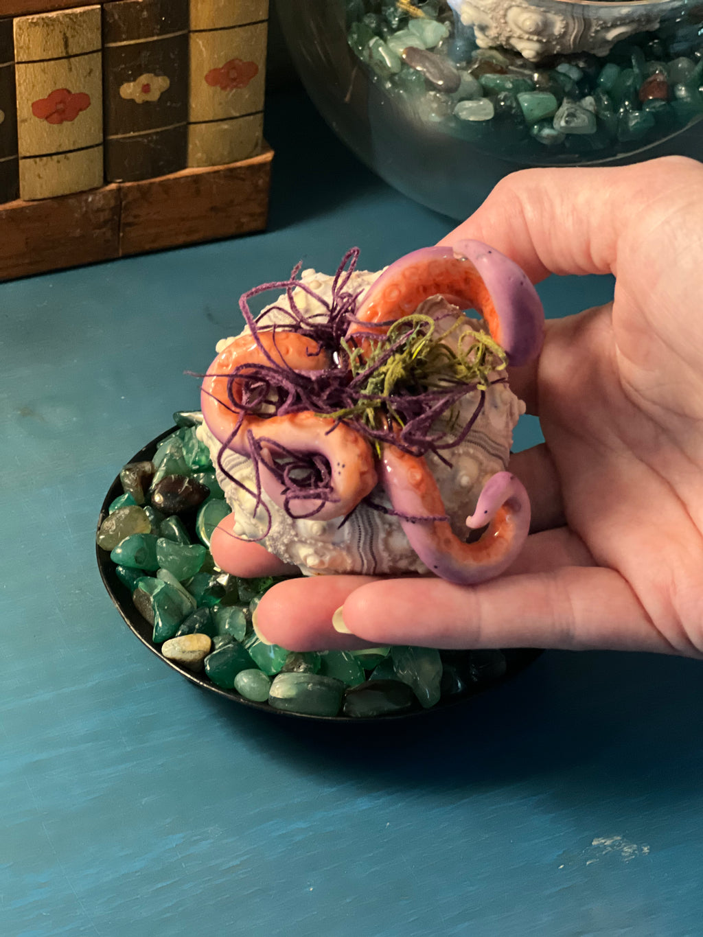 A hand holds a small decorative object made from a real clam shell, decorated with purple dried seaweed and green moss, set against a blue tabletop with green stones.