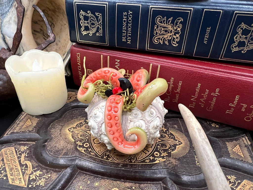 A decorative carved octopus figurine with pink tentacles, green accents, and a white textured body, styled on a dark ornate book with stacked antique-looking books and a candle in the background.