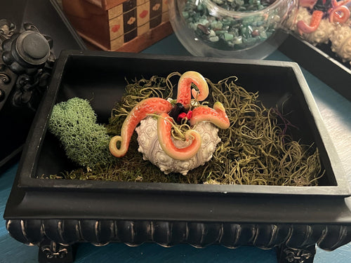 A decorative black rectangular tray filled with green moss, featuring a creature with tentacles, with a green mossy ball beside it.