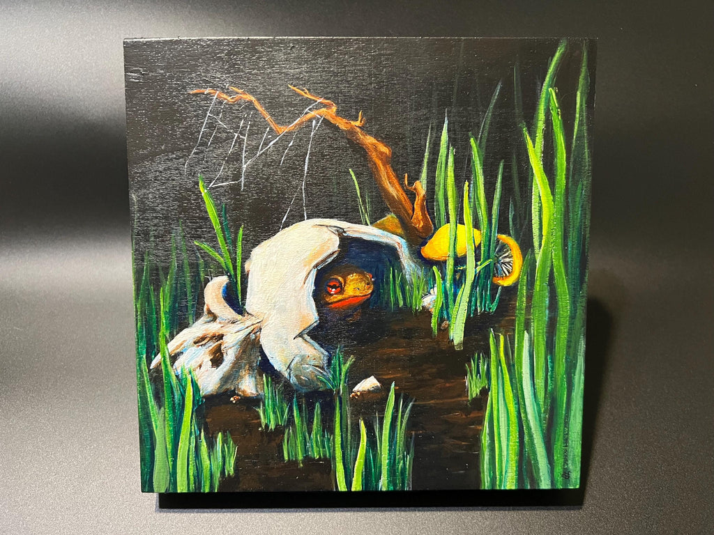 A square wooden painting depicting a surreal scene with a white skull, green reeds, yellow mushrooms, and a brown branch on a dark background, displayed against a neutral gray backdrop.