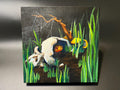 A square wooden painting depicting a surreal scene with a white skull, green reeds, yellow mushrooms, and a brown branch on a dark background, displayed against a neutral gray backdrop.