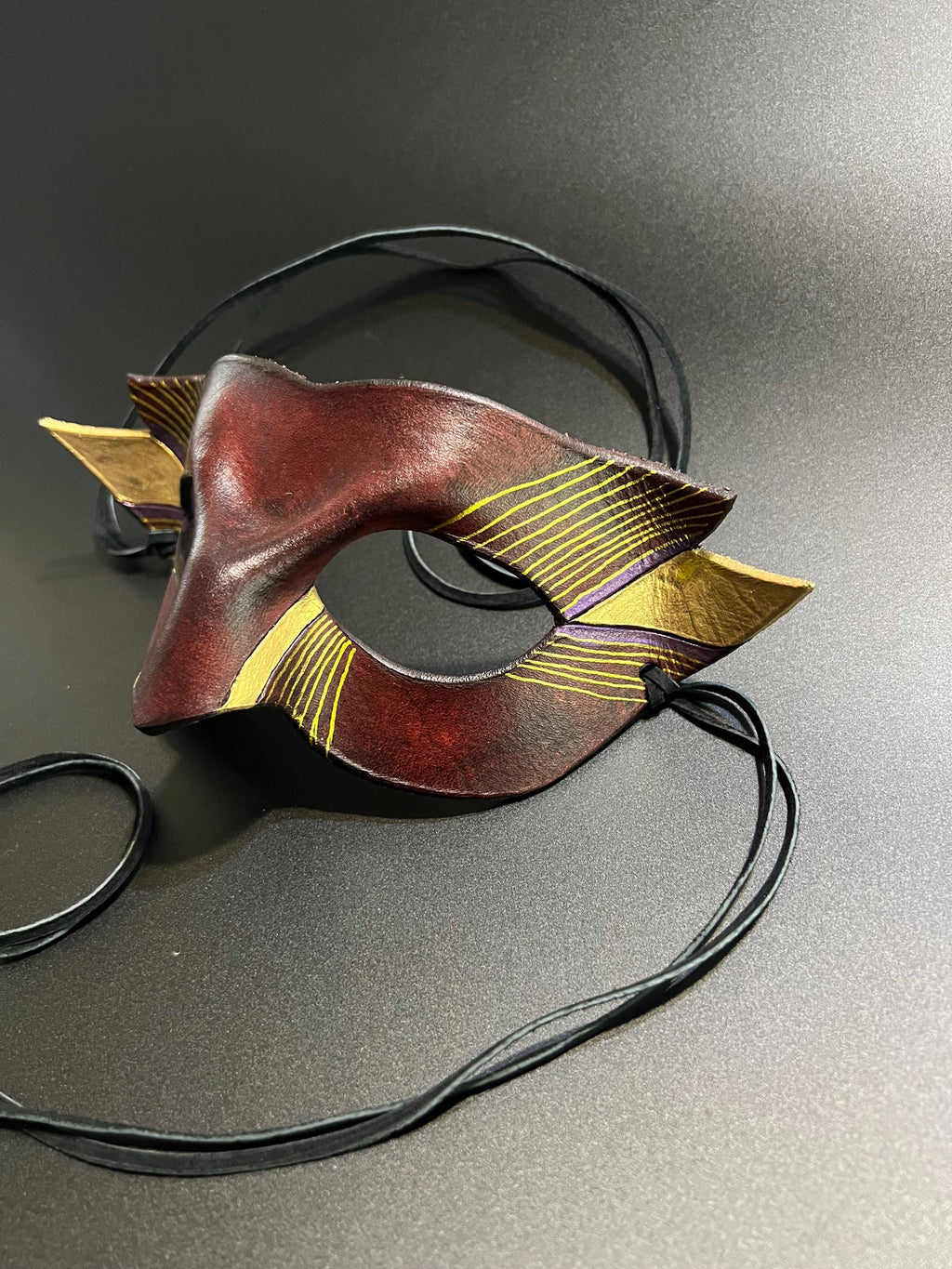 A decorative masquerade mask with a dark reddish-brown center, gold patterned side wings, and black suede ties, photographed on a gray studio background.