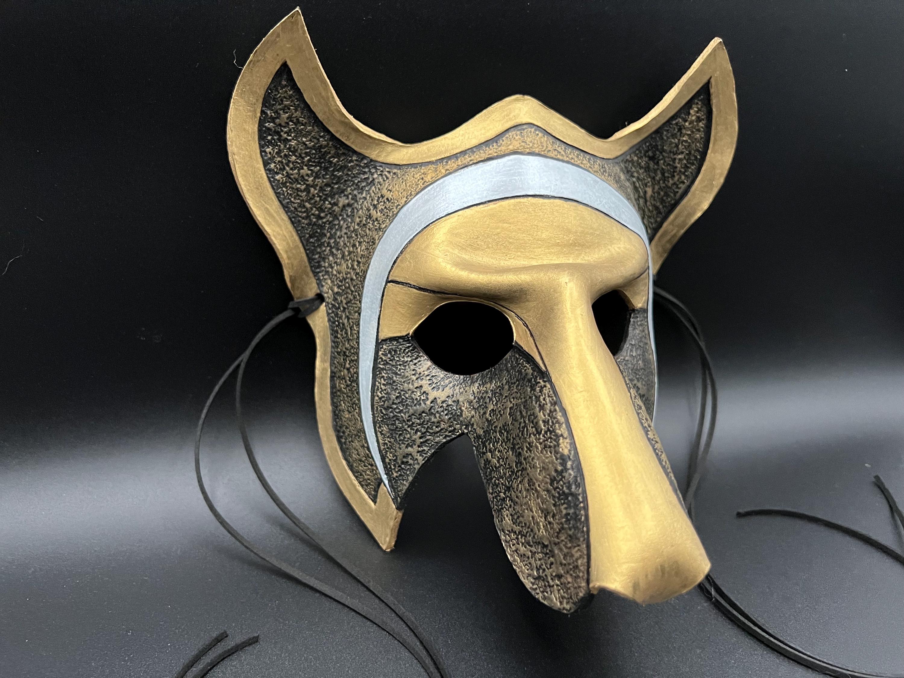 A stylized fox or wolf costume mask with a textured metallic gold and dark gray finish, featuring a silver band across the forehead and black hanging strings, shown against a dark background.