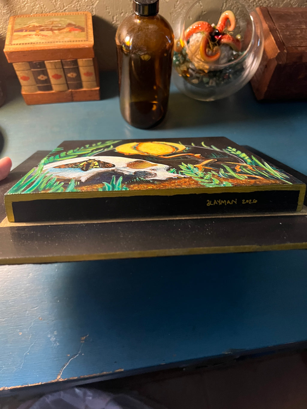 A hand-painted wooden plaque featuring a fantasy scene with a white skull, a yellow lamp, green grass, and a black background, resting on a blue wooden table.