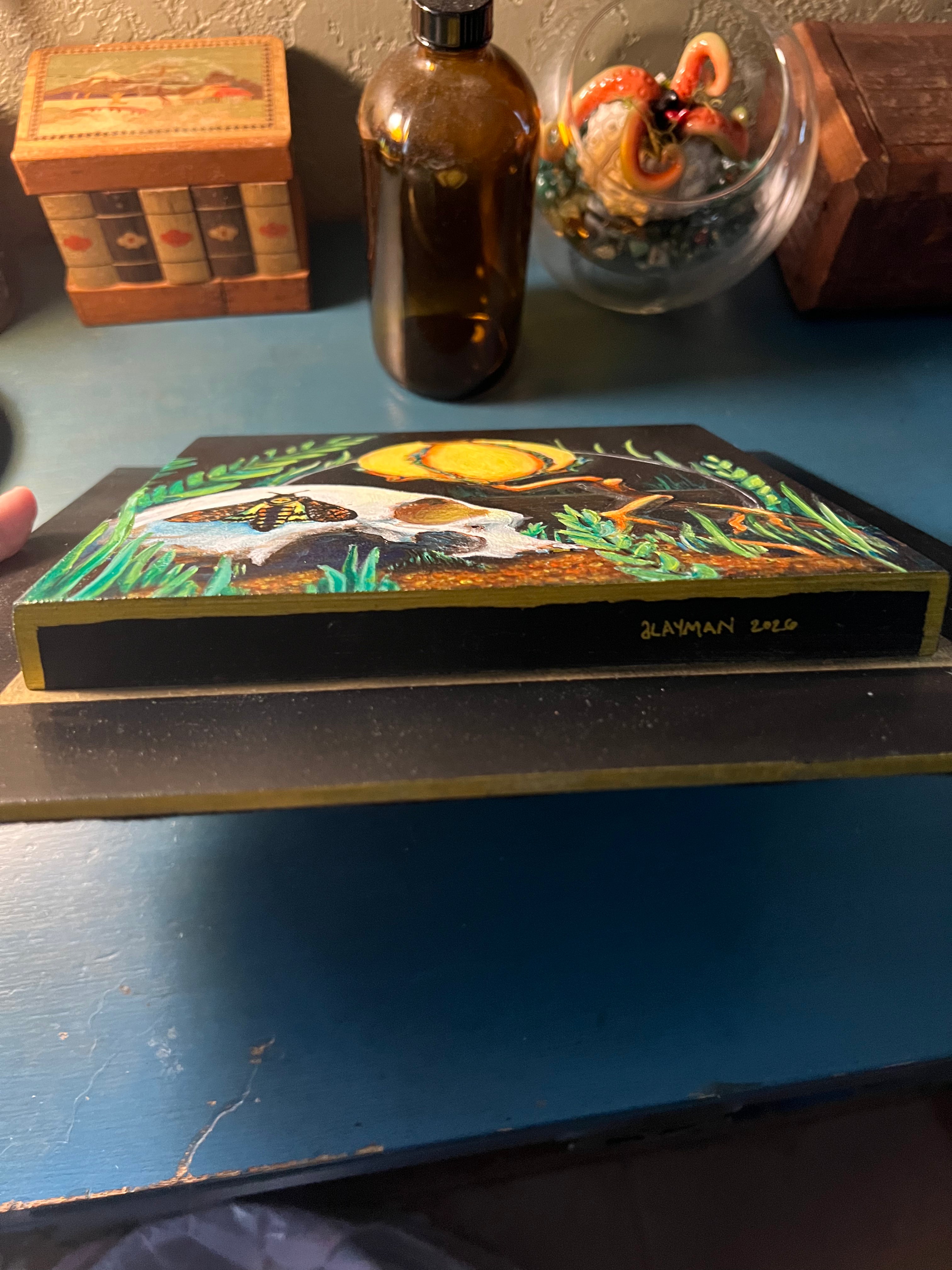 A hand-painted wooden plaque featuring a fantasy scene with a white skull, a yellow lamp, green grass, and a black background, resting on a blue wooden table.