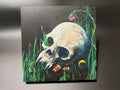 A square painted artwork showing a human skull nestled in tall green grass, with small colorful flowers and hidden coral snake. The painting is photographed against a dark gray wall.