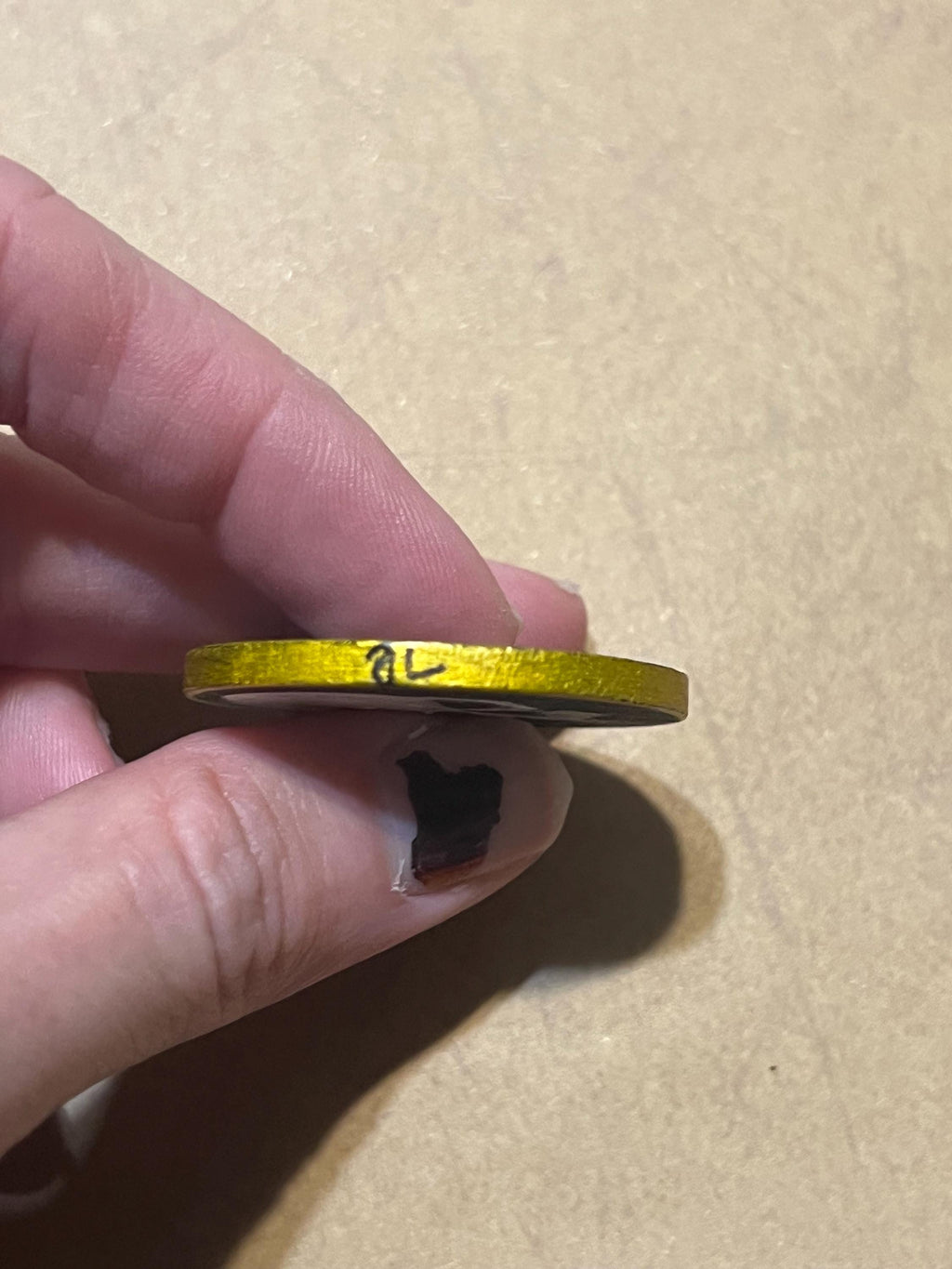 A hand holding a small gold-colored circular item, possibly a coin or ring, with handwritten markings on its top surface, against a plain tan background.