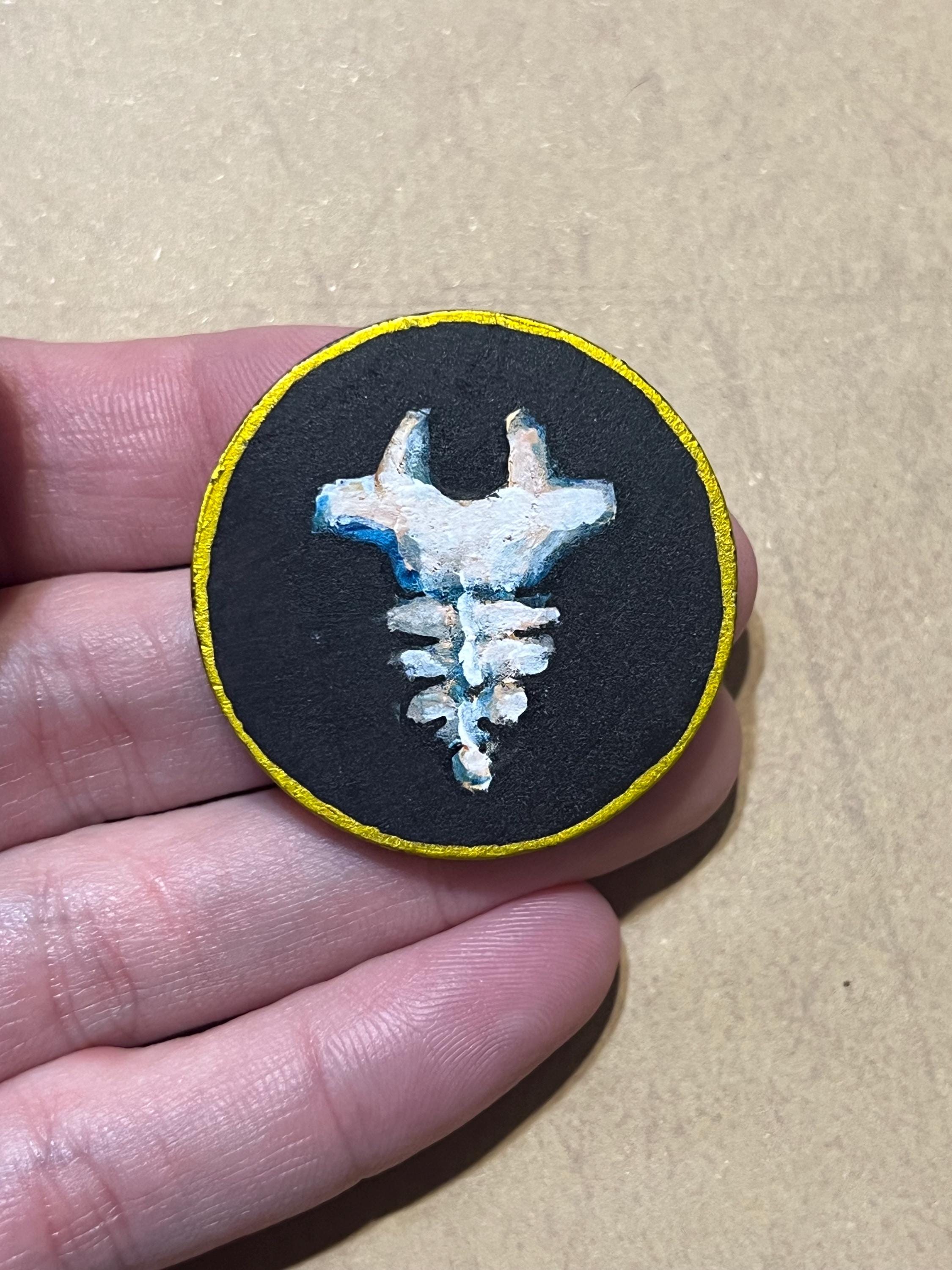 A hand holding a round black patch with a yellow border and a white and blue horned skull design in the center, set against a tan background.