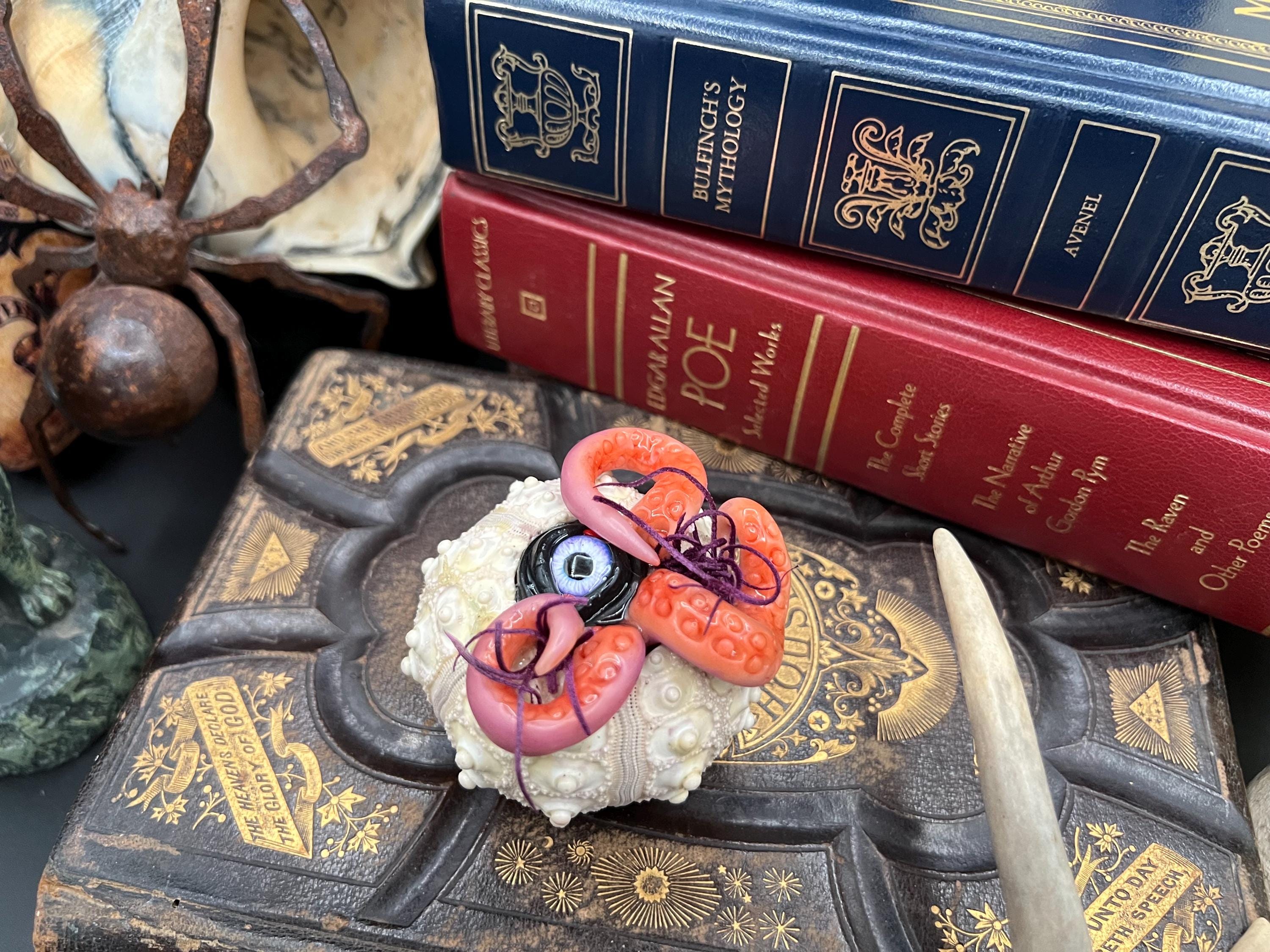 A small decorative object with a white textured base, pink and orange sculpted details, and a dark eye-like center, displayed on an ornate dark book with stacked blue and red books behind it.