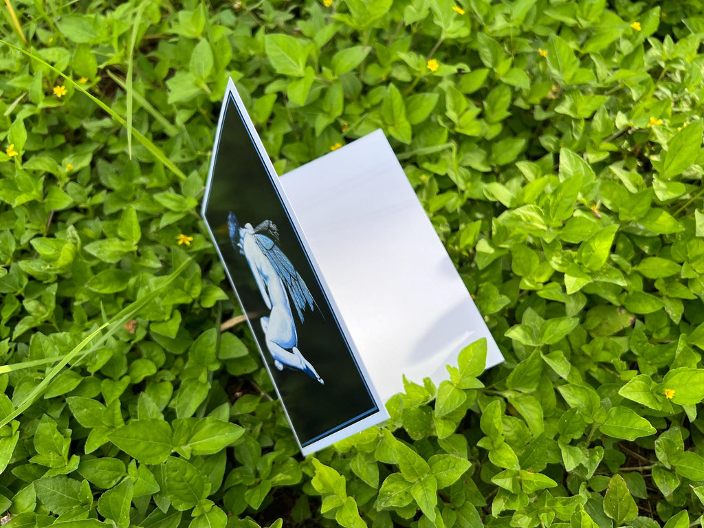 A small printed card or photo standing upright in dense green foliage. The card shows a black-and-white illustration of a winged figure.