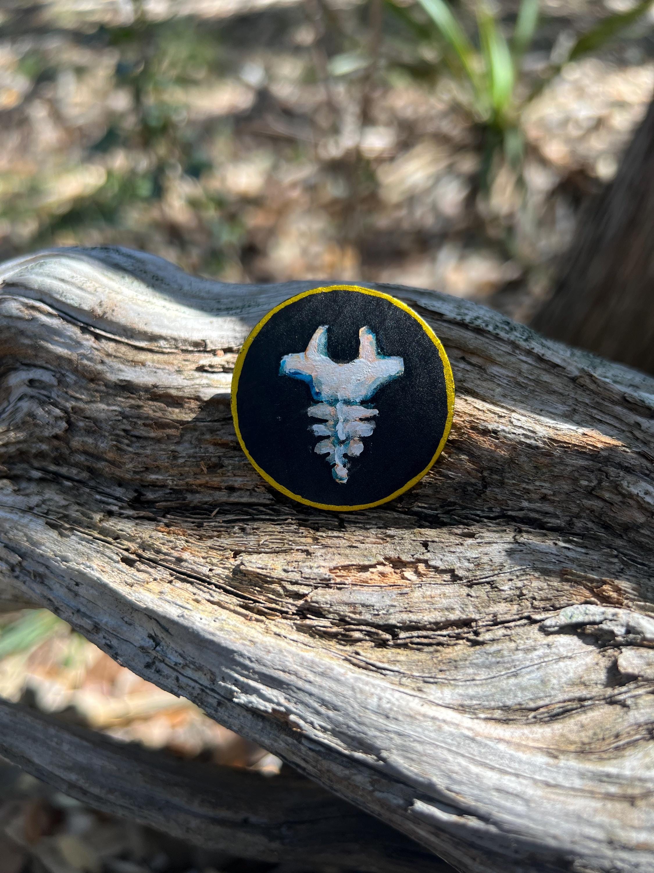 A round wooden token featuring a tail bone design with a gold border, resting on a weathered piece of wood outdoors.