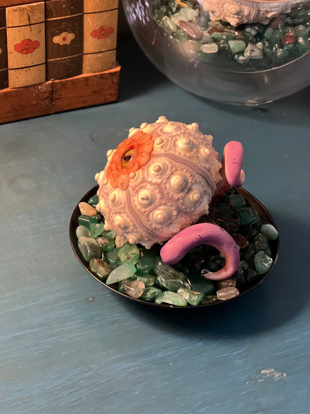 A decorative seashell-shaped ornament with pink tentacles sits in a small black dish filled with green and clear stones on a blue tabletop.