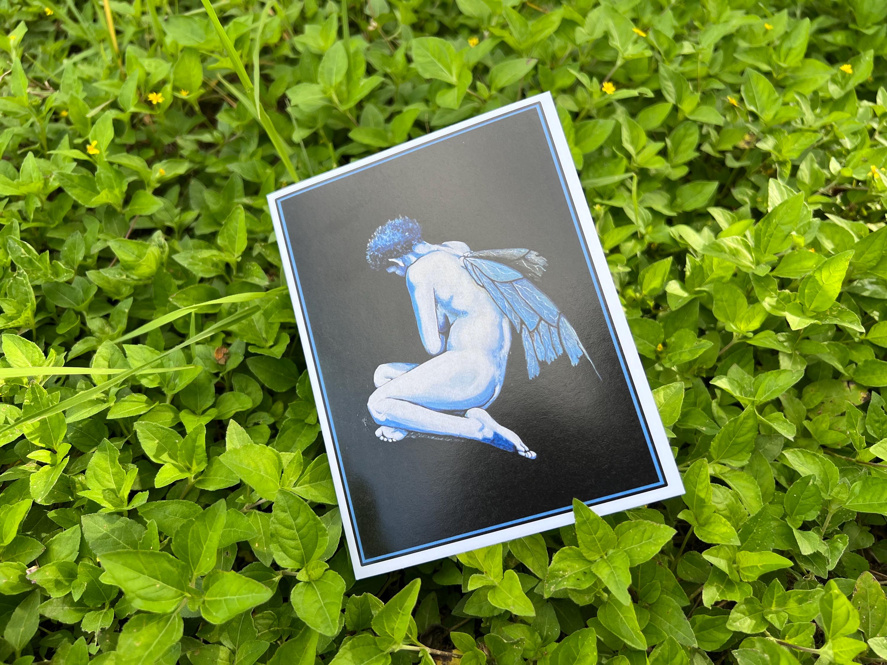 A rectangular art print or postcard featuring a pale blue winged nude figure on a dark background, placed on green leafy foliage outdoors.