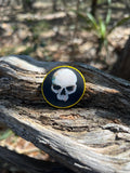 A round wooden token featuring a white skull design with a gold border, resting on a weathered piece of wood outdoors.