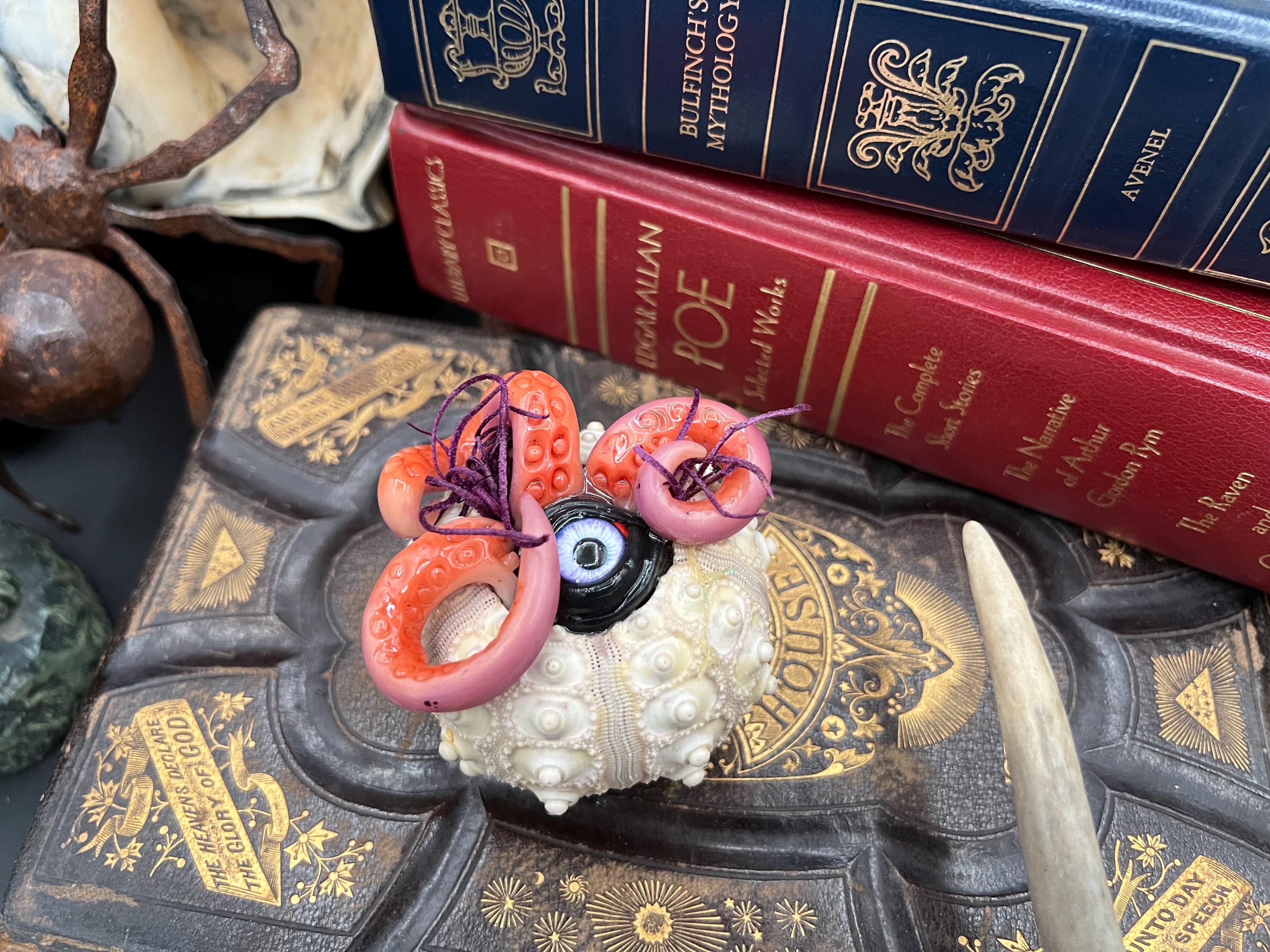 A small decorative object with a white textured base, pink tentacle-like loops, a black-and-white eye detail, and purple strands, displayed on top of an ornate dark book with gold detailing, with stacked blue and red books in the background.