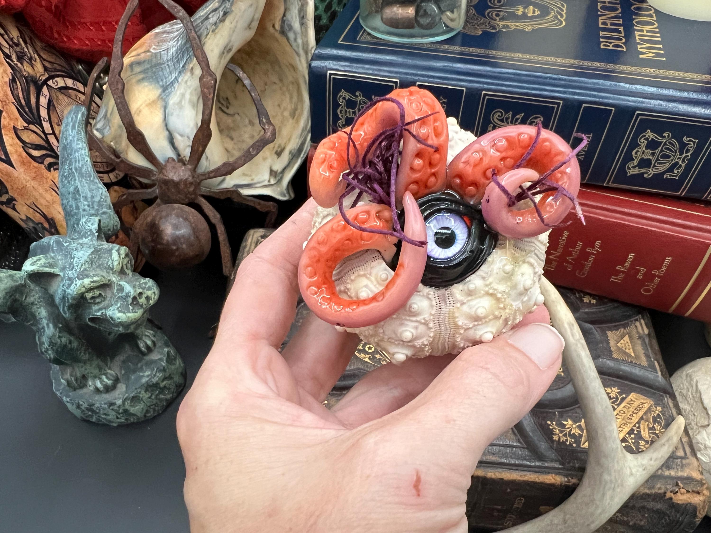 A hand holding a decorative carved skull object with coral-like orange and pink protrusions, photographed in a styled setting with books, a spider figurine, and other artifacts in the background.
