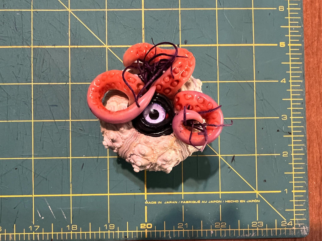 A small handmade horror prop resembling a severed eyeball with pink tentacles and a beige textured base, placed on a green cutting mat with yellow grid lines.