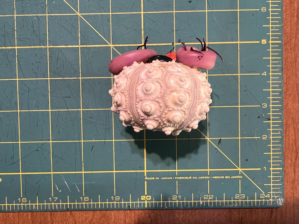 A small pink textured spherical item with two dark pink curved pieces on top, placed on a green cutting mat with yellow grid lines and a wooden surface around it.